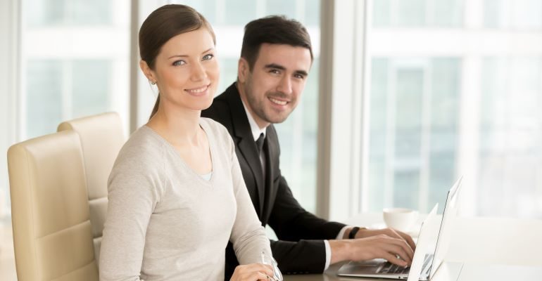 Two people in an office, sitting with laptops and smiling