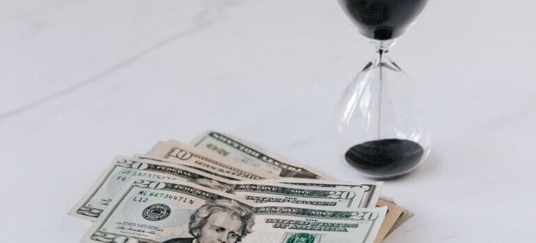 A stack of US dollar bills next to an hourglass on a white table.