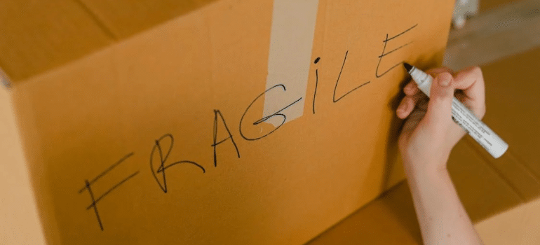 A close-up of a person writing the word “fragile” on the side of a moving box with a marker.