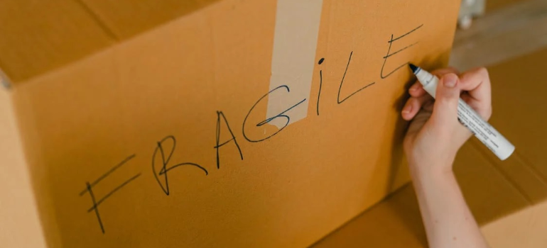 A close-up of a person writing the word “fragile” on the side of a moving box with a marker.