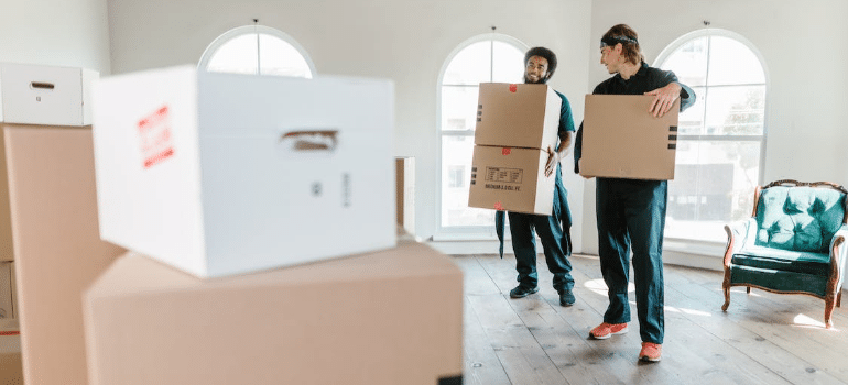 Two smiling movers carrying boxes toward a pile of boxes in an empty room.